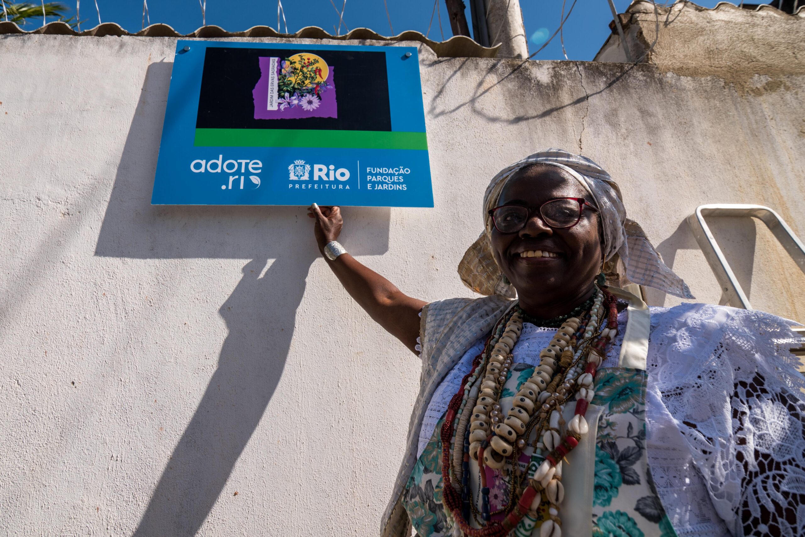 Luizinha de Nanã a frente de seu terreiro, segura placa da Prefeitura da cidade do Rio de Janeiro, que reconhece o Jardim das Ervas Sagradas como local de benefício prestado à sociedade. Foto: Bárbara Dias/RioOnWatch