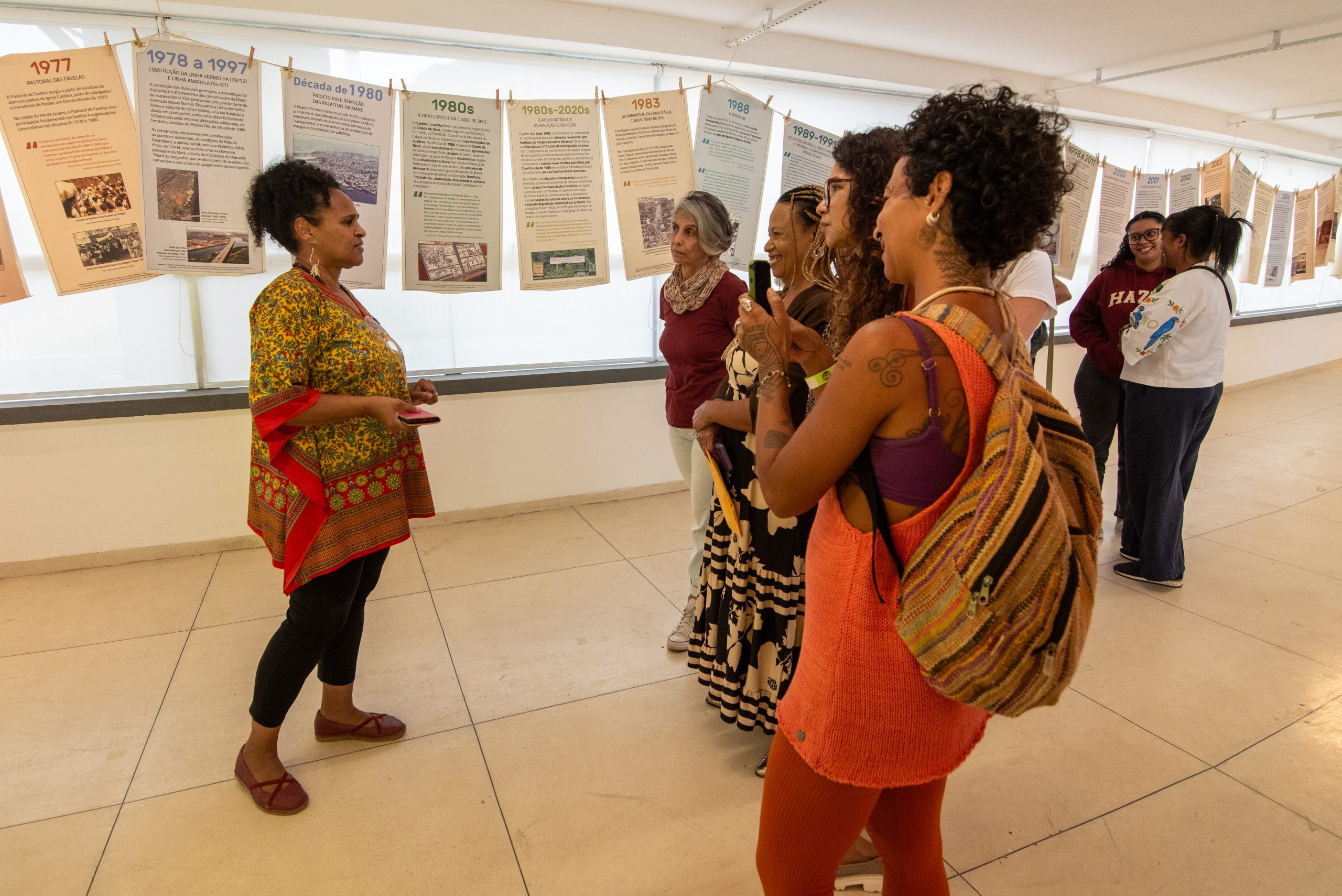 Márcia Souza (à esquerda) guia as visitantes, que durante o circuito da Exposição Memória Climática nas Favelas, onde puderam acompanhar os painéis da linha do tempo, o poço, a bacia das memórias e um vasto conteúdo sobre as memórias climáticas de dez favelas cariocas. Foto: Bárbara Dias Márcia Souza (à esquerda) guia as visitantes, que durante o circuito da Exposição Memória Climática nas Favelas, onde puderam acompanhar os painéis da linha do tempo, o poço, a bacia das memórias e um vasto conteúdo sobre as memórias climáticas de dez favelas cariocas. Foto: Bárbara Dias